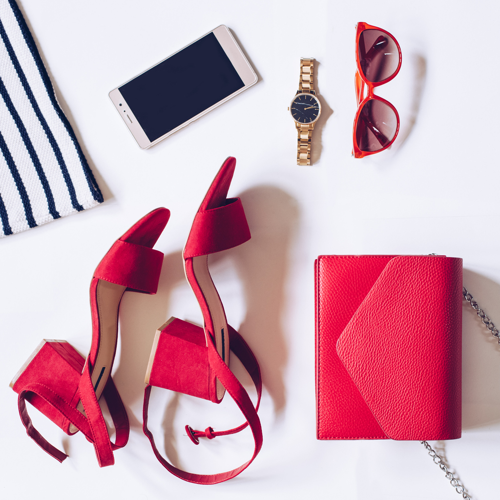 flat lay of a minimal set of female accessories:golden wrist watch, red mid heel sandals with ankle strap, clutch bag, mobile phone, sunglasses, striped shirt
